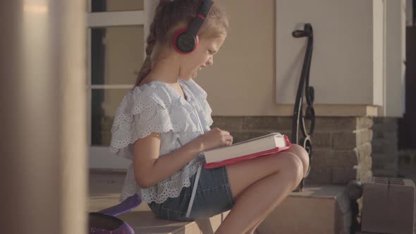 Adorable Girl During Summer Vacation Sitting on the Courtyard Near House Listening To Music, Singing