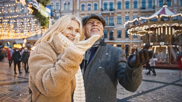 Happy Senior Couple Enjoying Snowfall Walking at Christmas Fair on City Square Slow Motion