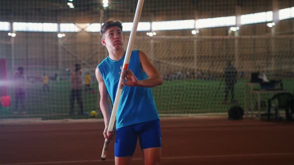Pole Vaulting Indoors - a Young Man in Blue Shirt Standing on the Track with a Pole and Preparing