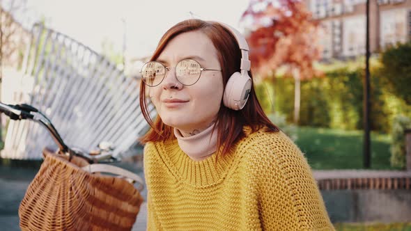 Young Woman Hipster Sitting on Bench in City Park Listening to Music Through Headphones Smiling