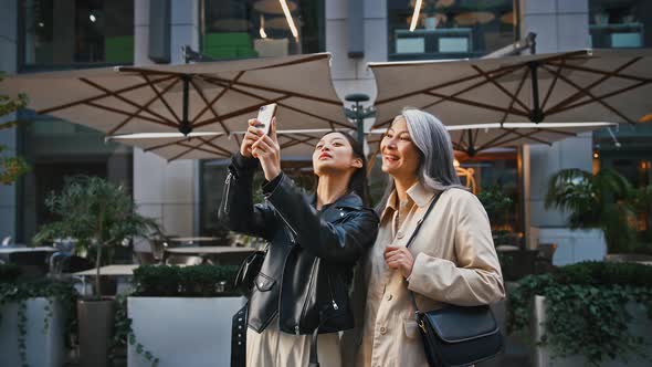 Asian Mature Woman and Her Adult Daughter Using Their Cellphones Taking a Selfie and Smiling While