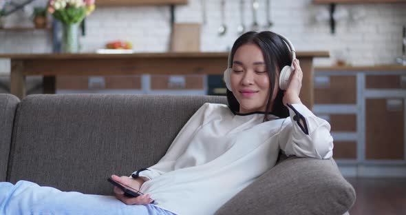 Asian Attractive Woman Listening to Music with Headphones Holding Smartphone on Sitting on Couch at