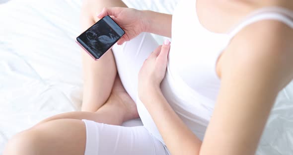 Pregnant Woman Sitting on Bed and Looking at Ultrasound Picture on Mobile Phone Closeup Movie Slow