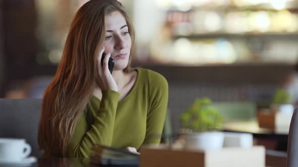 Attractive young woman is sitting in a cafe indoors and talking on the phone. Girl has a cup of coff