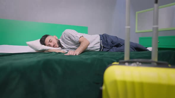 Wide Shot of Young Handsome Man Sleeping on Comfortable Cozy Bed with Packed Yellow Travel Bag at