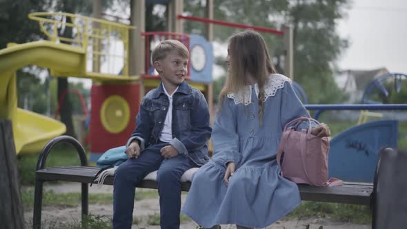 Two Relaxed Positive Children Sitting on Bench on Playground and Talking