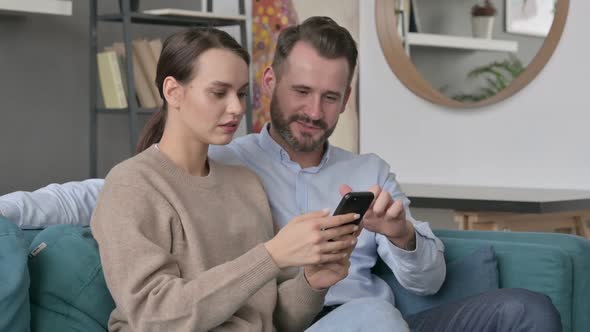 Portrait of Couple Celebrating Success on Smartphone Sitting on Sofa