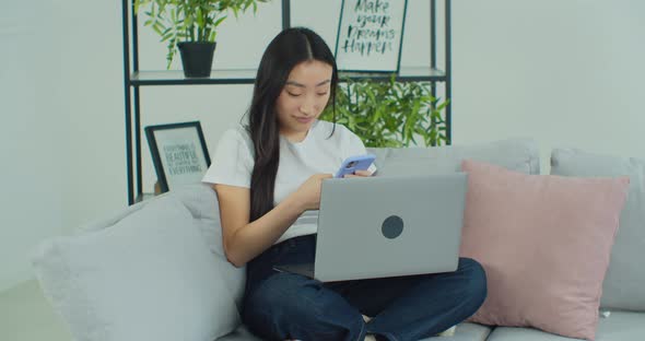 Woman Sitting in Front of the Laptop and Typing Messages on Her Smartphone While Sitting on a Couch