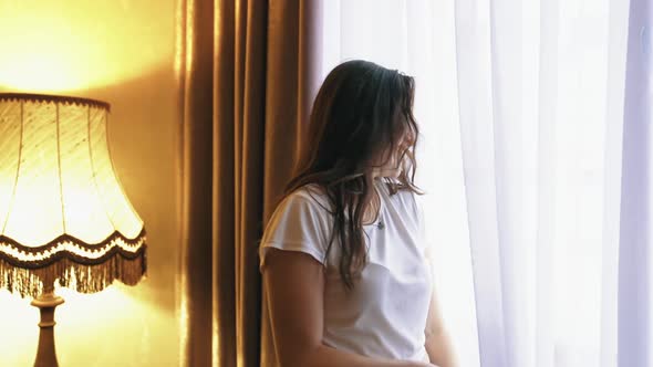 Young happy smiling woman looks out from a window of a hotel room. Large windows 