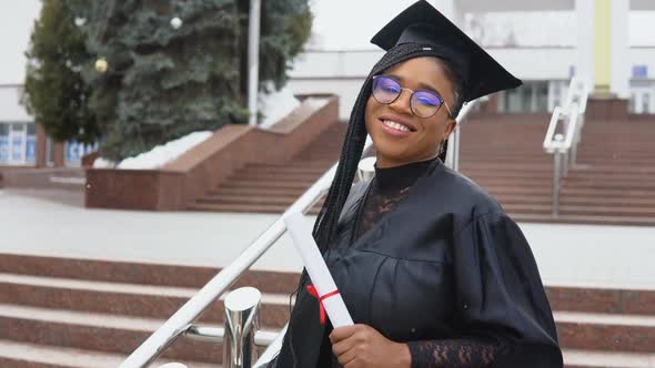 A Young Woman at the University at the Master's Mantle Holds a Diploma and Looks at the Camera