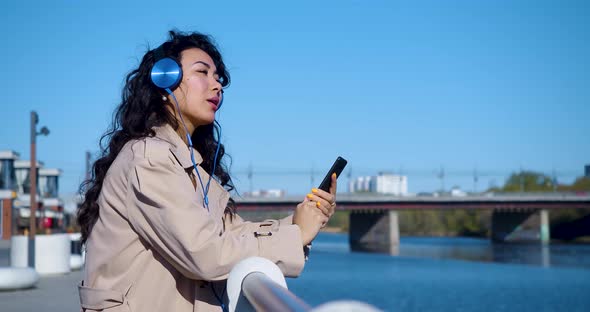 A Young Asian Woman in Headphones Listens to Music Through a Smartphone Application and Sings