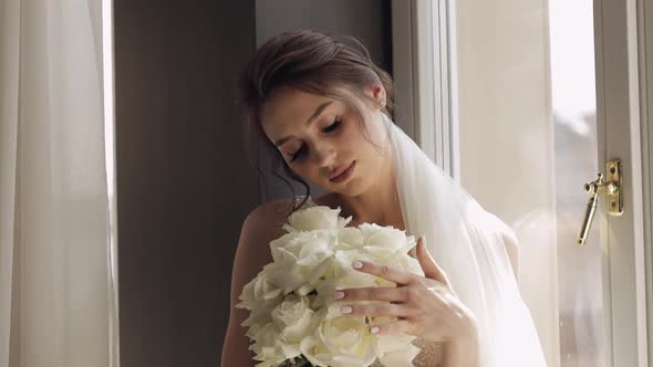 Bride in White Dress Staying Near Window with Flowers Bouquet Wedding Morning Preparations at Home