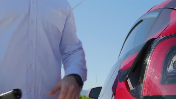 Man Connects an Electric Car to Charger at Wind Generators on Motion Background