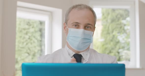 Portrait of Serious Caucasian Doctor in Face Mask Talking at Selfie Camera on Laptop and Gesturing