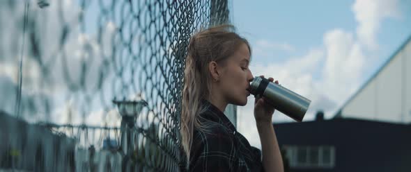 Young woman leaning towards metal fence and she drinks from bottle