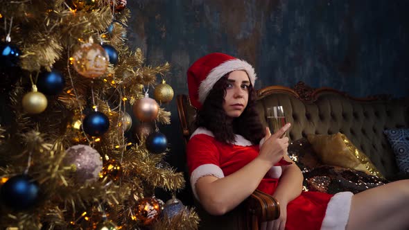 Young Woman in Santa Claus Costume with Glass of Champagne Sitting on Chair Near Christmas Tree