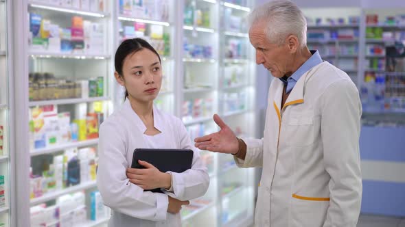 Experienced Caucasian Senior Man Scolding Young Asian Woman in Pharmacy