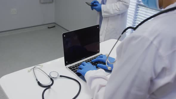 Midsection of doctor wearing face mask and surgical gloves using laptop, copy space on screen