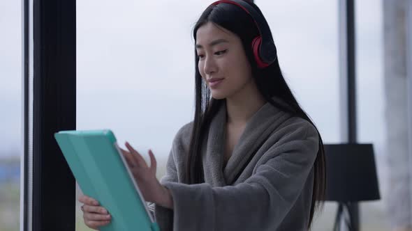 Young Relaxed Asian Woman in Headphones Surfing Social Media on Tablet at Window Indoors