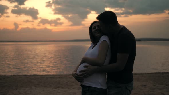 Husband Embraces His Beloved Pregnant Wife Standing On The Beach At Sunset