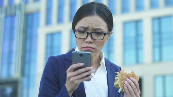 Young Female Manager Eating Unhealthy Burger in Hurry, Checking News on Phone