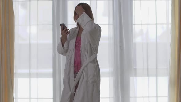 Plus Size Girl in White Long Bathrobe Dancing in Front of Large Floor-to-ceiling Window
