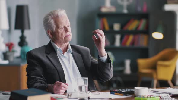 Elderly Man Taking Pill at Home, Sitting at Living Room Table