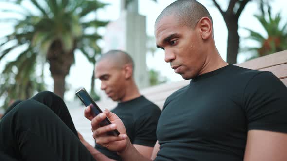 Two concentrated bald African brothers looking at phones while sitting on the bench