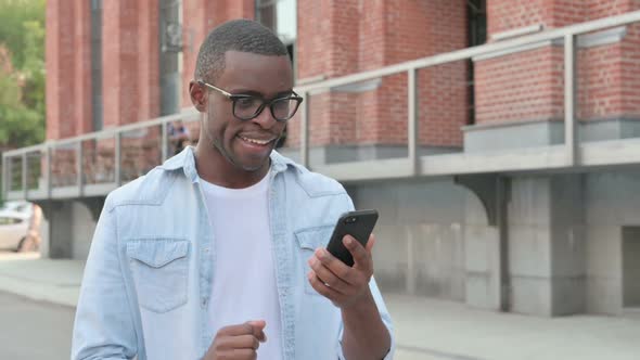 African Man Celebrating Success on Smartphone While Walking in Street