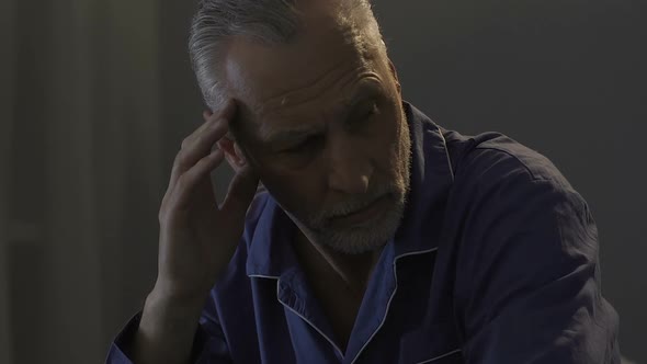 Elderly Male Sitting on Bed in Dark Room, Rubbing His Temples, Strong Headache