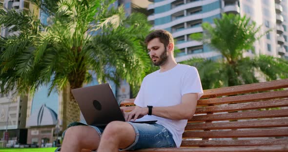 A Young Man in the Summer of Palm Trees Sitting with a Laptop and Typing on the Keyboard