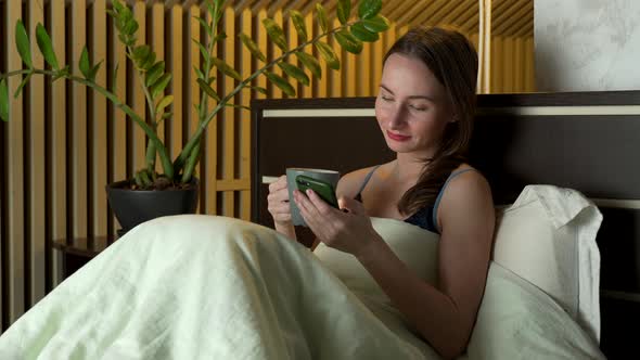 Woman Lies in Bed in the Evening and Uses a Smartphone and Drinks Tea