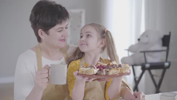 Happy Middle-aged Caucasian Woman Smiling at Camera and Hugging Little Pretty Girl with Pancakes on