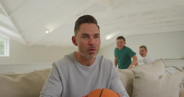Two caucasian boys hugging their father holding basketball and sitting on the couch at home