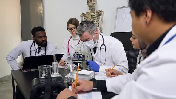 Group of Young Interracial Scientists Sitting at Work Table Working with a Microscope, Test Tubes