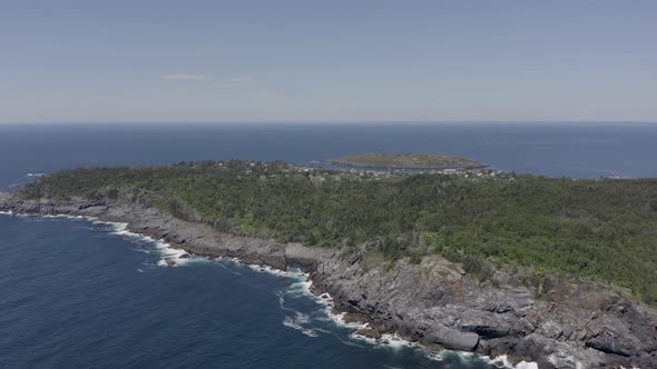 Drone shot flying above a small island in Maine with a rocky coastline and the town in the backgroun