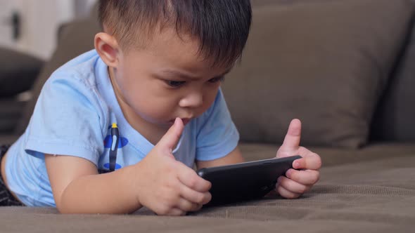 Asian Little Boy Laying On A Sofa Plays With Smartphone