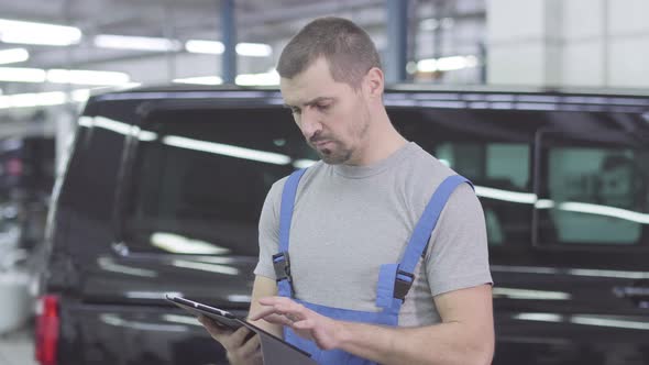 Portrait of Young Caucasian Auto Mechanic Typing on Tablet, Looking at Camera and Showing Thumb Up