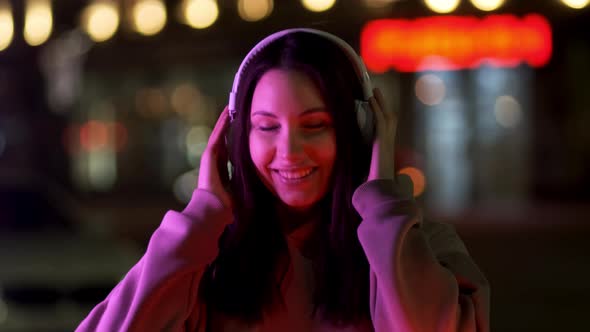 A Beautiful Young Woman with Glasses and an Earpiece Listens to Music on the Street at Night