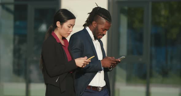 Asian Woman and Black Man with Phones Walk Along Street