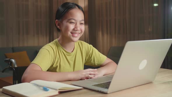 Asian Kid Girl Sitting In A Wheelchair And Raise Hand While Learning Online On Laptop Computer