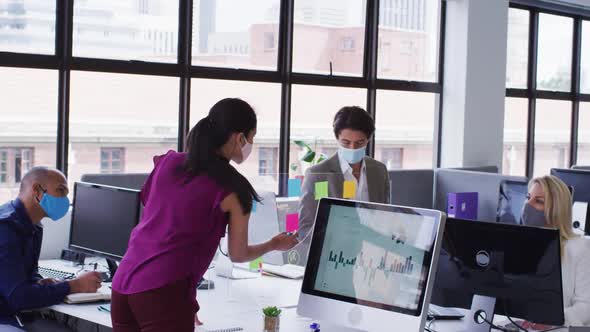 Diverse business colleagues wearing face masks using computers talking in office