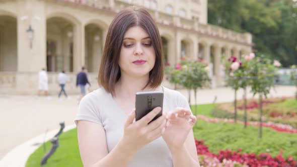 A Young Caucasian Woman Works on a Smartphone and Talks to the Camera in a City Park