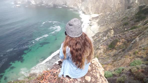 A young woman sitting on top of a cliff with the Bixby Creek Bridge in the background.