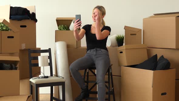 A Moving Woman Sits on a Chair in Empty Apartment and Takes Selfies, Surrounded By Cardboard Boxes