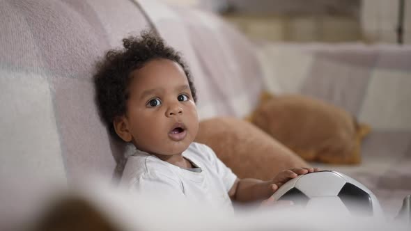 Adorable Little Cute African American Boy Sitting on Cozy Couch with Soccer Ball Looking Around