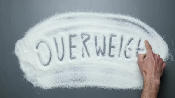 Male hand writes word overweight on sugar surface. No sugar. Sugar kills. Stop diabetes.