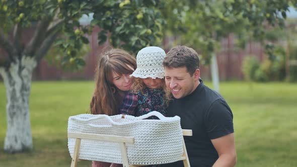 A Young Family Looks at Their Newborn Baby in a Cradle in the Garden