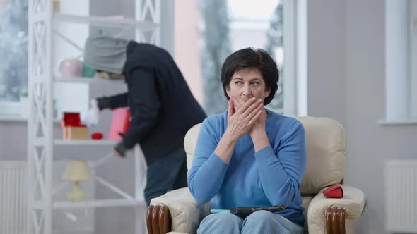 Frightened Nervous Woman Looking at Gun on Knees Closing Mouth with Hands As Blurred Robber