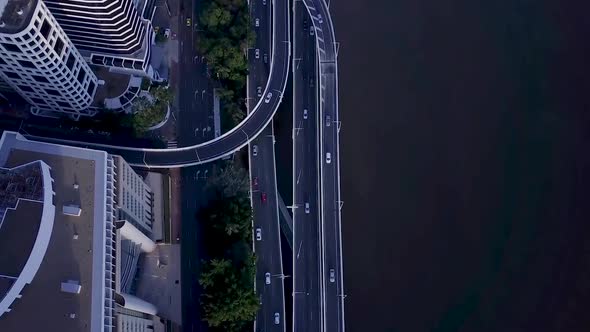 Aerial shot overhead of motorway with cars passing base of tall city building along river Brisbane,